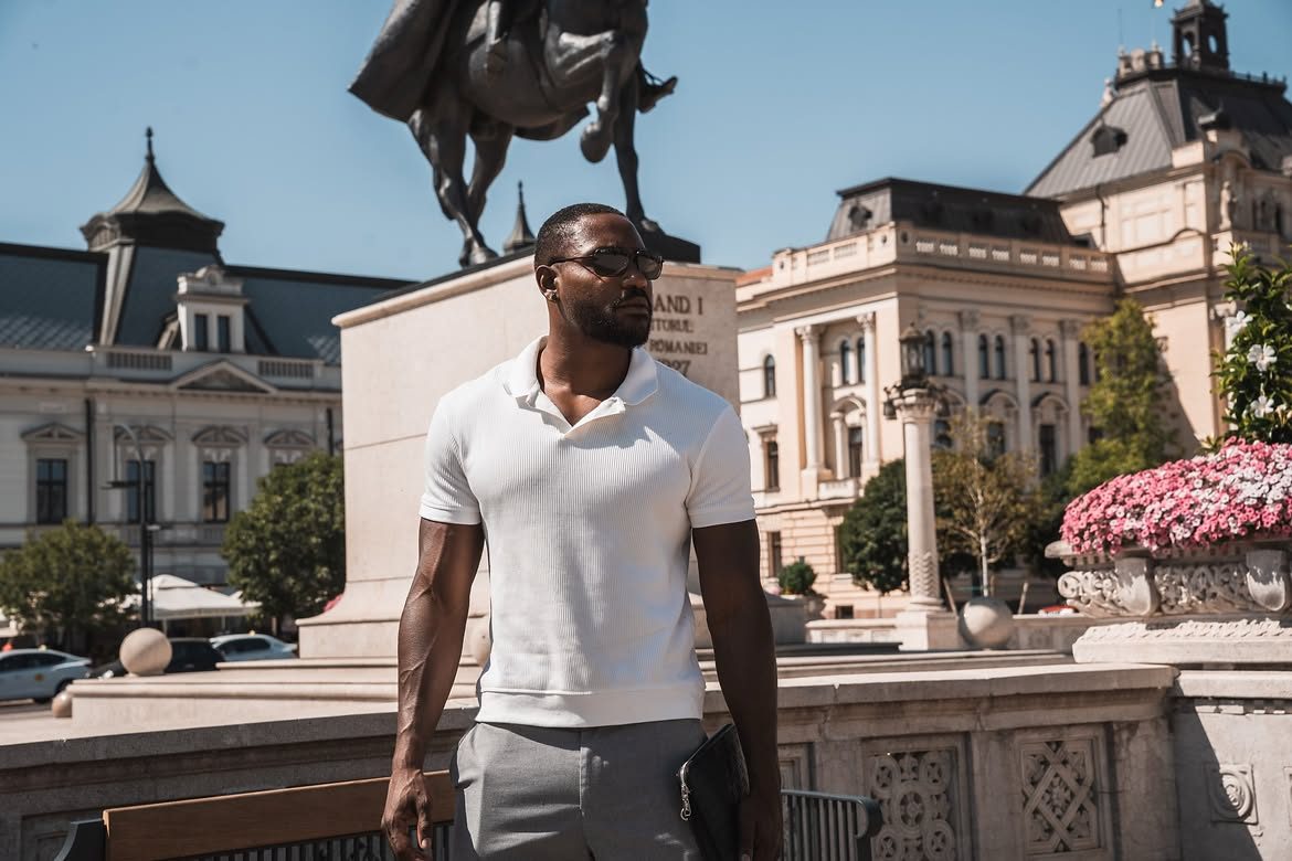 Martel Metellus in a white polo and sunglasses, standing in front of a monument in a European square